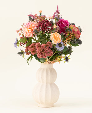 In einer weißen, geschwungenen Keramikvase befindet sich der Kunstblumenstrauß Pretty Pastel aus Seide – eine farbenfrohe Mischung aus nachhaltigen Blumen wie rosa Rosen, Dahlien, lila und orangefarbenen Blumen und Grün – wunderschön arrangiert vor einem hellen Hintergrund.