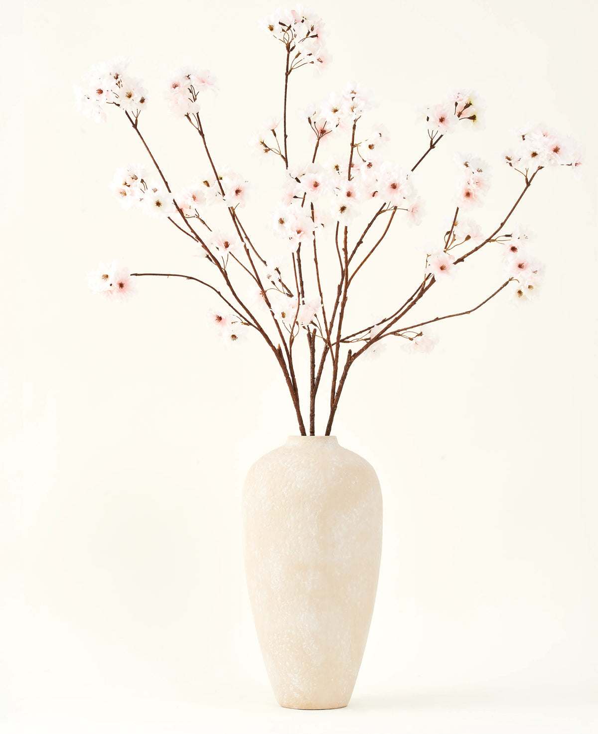 Der künstliche Seidenblumenstrauß Cherry Blossom Blush 114 cm hat hohe, zarte Zweige in einer schlichten beigen Vase, die Ihrem Interieur vor einem schlichten, hellen Hintergrund eine sanfte, elegante Note verleiht.