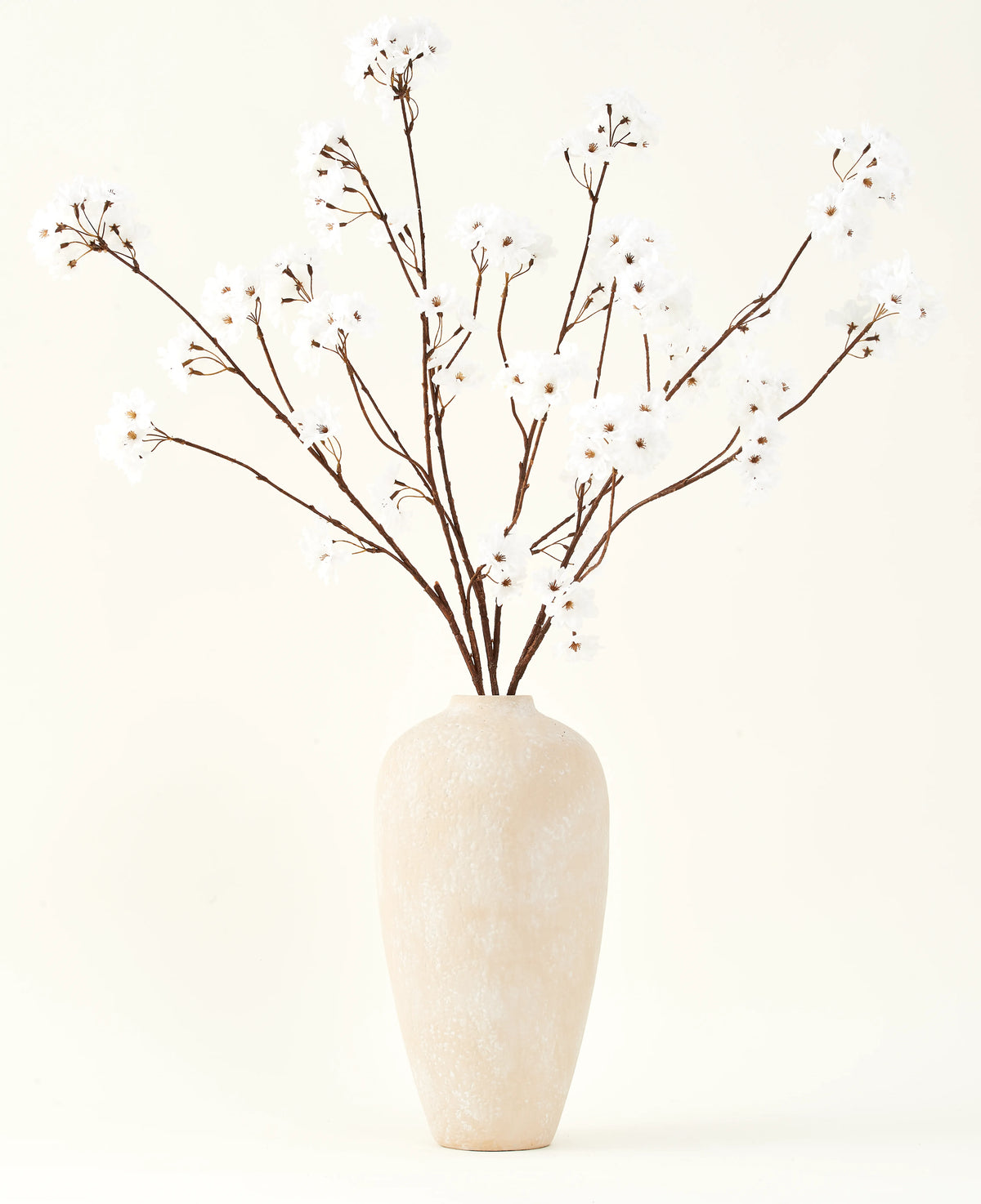 Der Seiden-Kunstblumenstrauß Kirschblütencreme 114 cm steht hoch in einer beigen Keramikvase und bringt mit seinen zarten, cremefarbenen Zweigen vor einem hellen Hintergrund eine heitere Dekoration in jeden Raum.