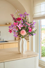 Auf einer Kücheninsel aus hellem Holz steht eine weiße Vase mit dem künstlichen Seidenblumenstrauß „Purple Dawn“ – bestehend aus rosa und lila Kunstblumen – mit einem Fenster und beigen Küchenschränken im Hintergrund.