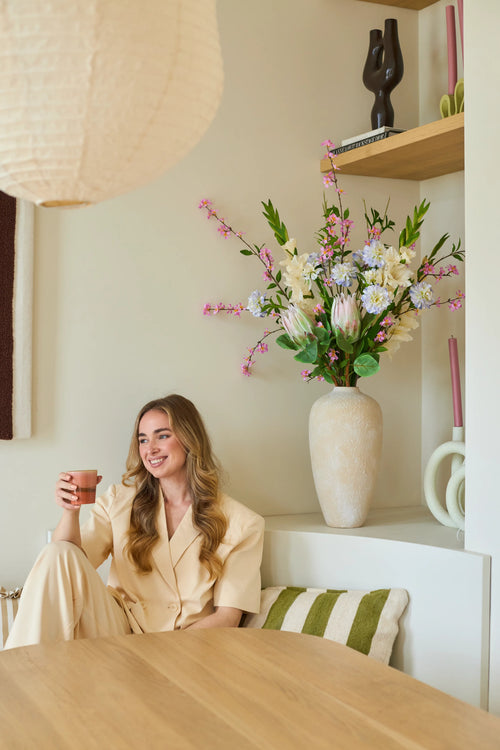 Eine Frau im cremefarbenen Anzug sitzt mit einer Tasse in der Hand und einem Lächeln im Gesicht auf einer Bank. Hinter ihr steht eine große Vase mit dem künstlichen Seidenblumenstrauß „Lots of Love XL“, bestehend aus rosa, weißen und gelben Blumen, mit Regalen im Hintergrund, auf denen Deko-Objekte ausgestellt sind. Das Ambiente wirkt gemütlich und modern.