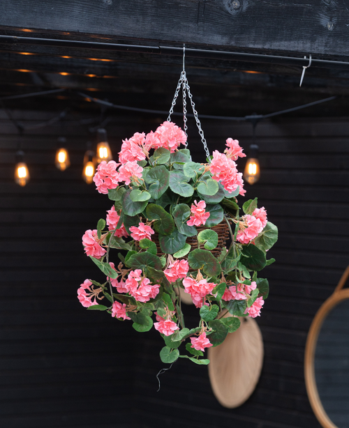Die künstliche Begonie rosa im Korb UV 70cm, mit sattgrünen Blättern und rosa Blüten, hängt an einer Kette. Warmes Streiflicht und ein runder Spiegel betonen den dunklen Holzhintergrund.