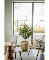 Ein künstlicher Orangenbaum 120 cm steht neben einem großen Fenster in einem modernen, hellen Wohnzimmer mit beigem Stuhl, Beistelltisch aus Holz und einer Kaffeetasse auf einer Untertasse.