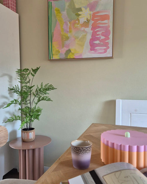 In einem pastellfarbenen Raum gibt es eine 85 cm große künstliche Farnpflanze auf einem rosa Beistelltisch, eine lila-goldene Tasse, eine rosa Schale mit Wellenrand, ein offenes Notizbuch und an der Wand ein abstraktes Gemälde mit Grün-, Rosa- und Gelbtönen.