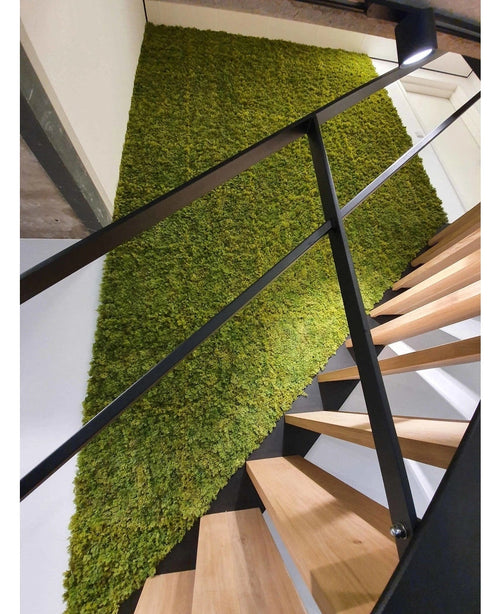 Eine moderne Treppe mit Holzstufen und schwarzem Geländer steht neben einer grünen Wand aus Kunsthaag Nordic Moss 50x50 cm UV und setzt einen natürlichen, strukturierten Akzent in der luxuriösen Inneneinrichtung.