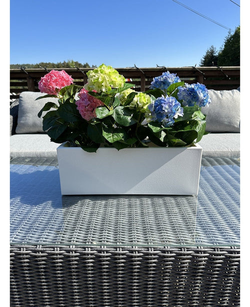 Die Kunstpflanze Hortensie Pink UV 35 cm, ein rechteckiger weißer Übertopf mit rosa Hortensien, steht draußen auf einem Korbtisch mit Glasplatte, grauen Kissen und einem blauen Himmel im Hintergrund.