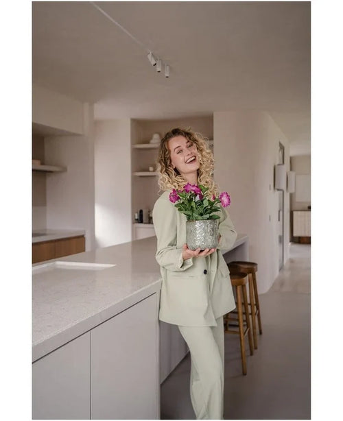 Eine lächelnde Frau mit lockigem Haar und hellem Anzug steht in einer modernen, minimalistischen Küche und hält eine künstliche Petunie UV 25 cm dunkelrosa mit leuchtend rosa Blüten in der Hand.