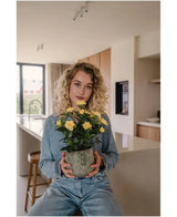 Eine Frau mit lockigen blonden Haaren, gekleidet in Jeanskleidung, sitzt mit der Kunstpflanze Mini Rose UV 35 cm gelb in den Händen in einer modernen Küche und blickt direkt in die Kamera.