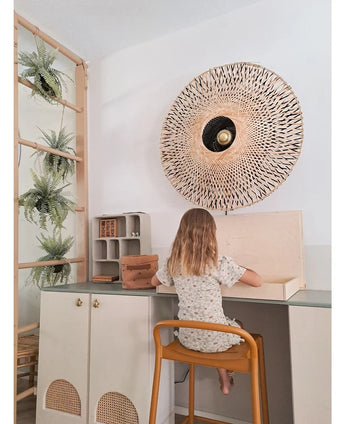 Ein kleines Kind mit schulterlangen Haaren sitzt an einem Schreibtisch in einem hellen Raum, den Rücken dem Betrachter zugewandt. Hinter ihr stehen eine geflochtene Wandlampe, einige Schränke und die künstliche Hängepflanze „Frosted Fern on ball 76 cm“, eingebettet zwischen den Pflanzen auf einer Holzleiter, daneben kleine Regale mit ordentlich angeordneten Gegenständen.