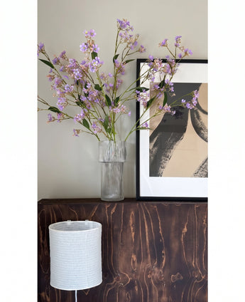 In einer transparenten Vase stehen die Blüten des Kunsttak Daphne Spray 108 cm lila auf einer dunklen Holzoberfläche neben einem modernen, abstrakten Kunstwerk in einem Rahmen, darunter ein weißer strukturierter Lampenschirm.