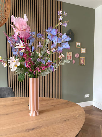 Eine hohe rosa-weiß gestreifte Vase mit dem künstlichen Seidenblumenstrauß Amethyst Romance steht auf einem runden Holztisch in einem modernen Raum mit grünen Wänden, Holzvertäfelung und kleinen gerahmten Kunstwerken.