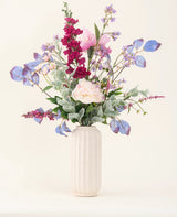 Eine weiße, strukturierte Vase präsentiert den „Silk Artificial Flowers Bouquet Amethyst Romance“ mit rosa Pfingstrosen, lila und blauen Blumen, burgunderfarbenen Blumen und grünem Blattwerk vor einem hellen, neutralen Hintergrund.