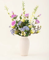 In een crèmekleurige vaas staat een Zijden kunstbloemen boeket Magnolia Glow met paarse, witte en gele bloemen-delphiniums, lisianthus, lelies en vergeet-me-nietjes tegen een effen lichte achtergrond.