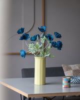 Eine 73 cm große künstliche Mohnblume in Dunkelblau steht in einer gelben Vase auf einem Tisch, begleitet von einer kleinen, bunt gestreiften Tasse, in einem modernen, sanft beleuchteten Raum mit minimalistischem Dekor und neutralen Tönen.
