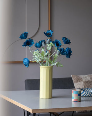 Eine 73 cm große künstliche Mohnblume in Dunkelblau steht in einer gelben Vase auf einem Tisch, begleitet von einer kleinen, bunt gestreiften Tasse, in einem modernen, sanft beleuchteten Raum mit minimalistischem Dekor und neutralen Tönen.