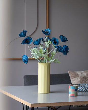 Eine 73 cm große künstliche Mohnblume in Dunkelblau steht in einer gelben Vase auf einem Tisch, begleitet von einer kleinen, bunt gestreiften Tasse, in einem modernen, sanft beleuchteten Raum mit minimalistischem Dekor und neutralen Tönen.