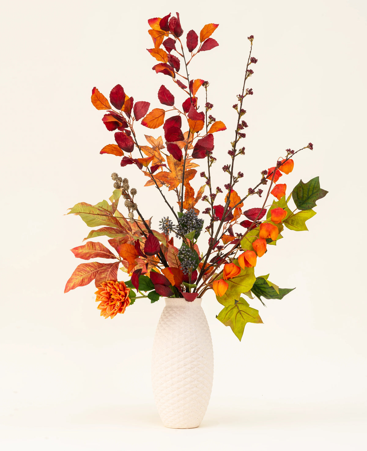 Eine weiße, strukturierte Vase präsentiert den künstlichen Seidenblumenstrauß „Chestnut Charm“ mit Herbstblättern, orangefarbenen Blüten und grünem Blattwerk – ein elegantes künstliches Arrangement vor einem schlichten, hellen Hintergrund.