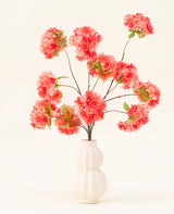 Het Zijden kunstbloemen boeket Kersenbloesem Koraal heeft levendige roze en koraal kunstmatige kersenbloesems met groene bladeren, elegant gerangschikt in een wit gebeeldhouwde vaas tegen een lichte achtergrond.