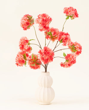 Het Zijden kunstbloemen boeket Kersenbloesem Koraal heeft levendige roze en koraal kunstmatige kersenbloesems met groene bladeren, elegant gerangschikt in een wit gebeeldhouwde vaas tegen een lichte achtergrond.