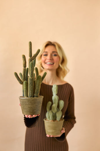 Eine lächelnde Frau in einem braunen Kleid hält zwei künstliche Kaktus-Cereus-Topfpflanzen (35 cm), eine große und eine kleine, vor einem schlichten beigen Hintergrund.