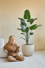 Een vrouw in een bruine jumpsuit zit glimlachend op de grond met een koffiekopje naast zich, naast de Kunstplant Strelitzia 180 cm real touch in een minimalistische kamer met beige muren.