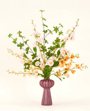 Een Zijden kunstbloemen boeket Blossom Harmony is gearrangeerd in een mauve vaas met roze en perzik bloemen en groene bladeren, tegen een eenvoudige lichte achtergrond voor een elegante display.