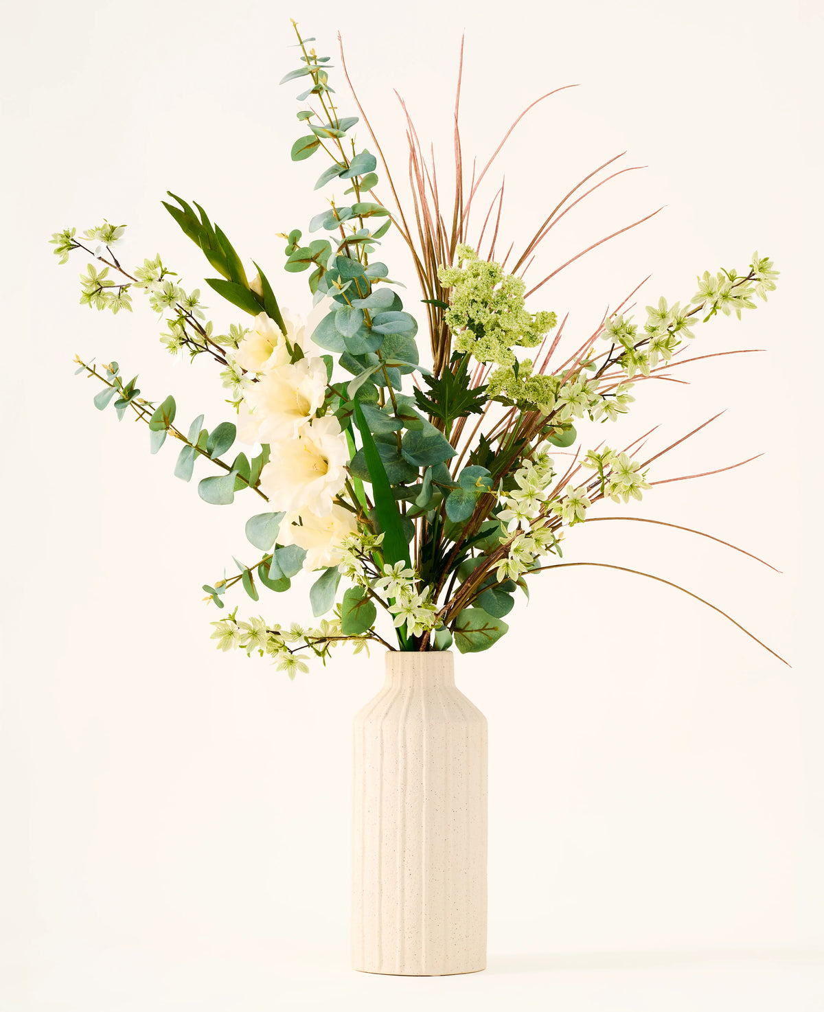 In einer beigen Vase befindet sich der Strauß „Wild Ivory“ aus künstlichen Seidenblumen mit grünen Blättern, Eukalyptus, weißen Blüten, kleinen grünen Blüten und langen, dünnen braunen Gräsern vor hellem Hintergrund.