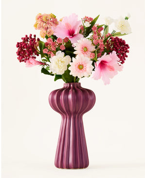 Een boeket kunstbloemen van Zijden, ‘Bloom & Berry’, met roze, witte en rode kunstbloemen, is prachtig geschikt in een hoge, geribbelde, paarse vaas tegen een effen witte achtergrond.