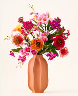 In einer pfirsichfarbenen Vase befindet sich der Strauß „Tropical Sunset“ aus Seiden-Kunstblumen, bestehend aus einem lebhaften Mix aus rosa, orange, gelben und roten Blüten vor einem schlichten, hellen Hintergrund.