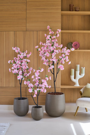 Drie Kunst Kersenbloesem-bomen van 120 cm, versierd met delicate roze bloesems, staan voor een houten lambrisering, naast een beige fauteuil en een moderne witte kandelaar op de plank. Deze elegante opstelling van kunstbloemen geeft elke ruimte blijvende schoonheid en verfijning.