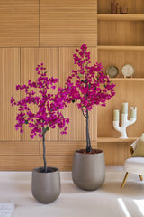Zwei Topfpflanzen vom Typ Künstliche Bougainvillea, 180 cm, in der Farbe Lila, mit leuchtend violetten Blüten stehen vor einer hellen Holzwand und Regalen mit Deko-Objekten wie Kerzen und Tellern. Auf der rechten Seite steht ein beigefarbener Stuhl, der diesem lebhaften Wohnzimmer mit künstlichen Pflanzen eine gemütliche Atmosphäre verleiht.