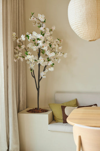 Eine 180 cm große künstliche Kirschblüte in Weiß steht neben einer Sitzecke mit Kissen in Grün- und Brauntönen, neben einem runden Holztisch und einer Papierlaterne in einem sanft beleuchteten Raum mit neutralen Farbtönen.