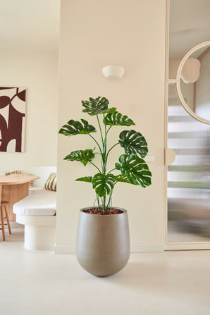 De Kunstplant Monstera van 120 cm staat in een moderne, minimalistische woonruimte met neutrale muren, een ronde spiegel, houten meubels en abstracte wandkunst.