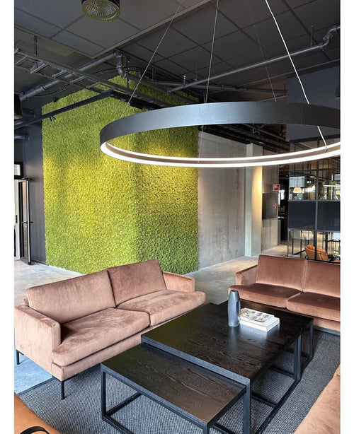 Eine moderne Sitzecke mit beigen Sofas, schwarzen Couchtischen, einer großen runden Deckenlampe und einer grünen Wand im Hintergrund, gestaltet mit Kunsthaag Nordic Mos 50x50 cm UV, die ein luxuriöses Interieur und ein zeitgenössisches Industriedesign ausstrahlt.