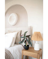 Een gezellige slaapkamer met neutrale tinten heeft een bed met wit linnen, een houten nachtkastje met de Kunst Begonia 48 cm in sierpot zwart en een geweven lamp, en ronde muurkunst met textuur boven het bed.