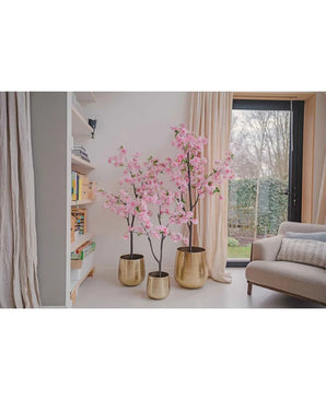 Drie Kunst Kersenbloesem 120 cm roze bomen in gouden potten staan naast een boekenplank en een raam in een lichte, gezellige woonkamer met beige gordijnen, een grijze bank en uitzicht op de tuin.