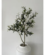 De Kunst Olijfboom 120 cm heeft groene bladeren en zwarte olijven in een witte pot met structuur en kiezels, waardoor het een prachtig decoratief element is tegen een effen lichtgrijze muur.