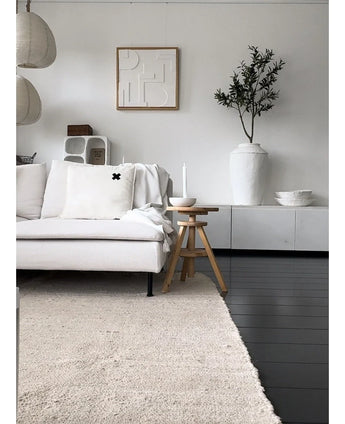 Een minimalistische woonkamer met een lichtgekleurde bank, neutraal vloerkleed, wit dressoir, Kunst Olijfboom 120 cm als decoratief element, abstracte muurkunst en houten krukjes-alles in zachte Mediterrane sfeer met neutrale tinten.