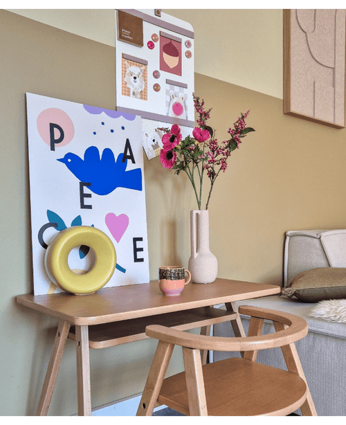 Ein Holzschreibtisch mit passendem Stuhl zeigt einen blauen Vogelaufdruck, eine künstliche Blume Anemone verzweigt 56 cm rosa, ein rundes gelbes Ornament und eine kleine Tasse. Oben hängt neben einem beigen Sofa eine Pinnwand mit Fotos und Notizen.