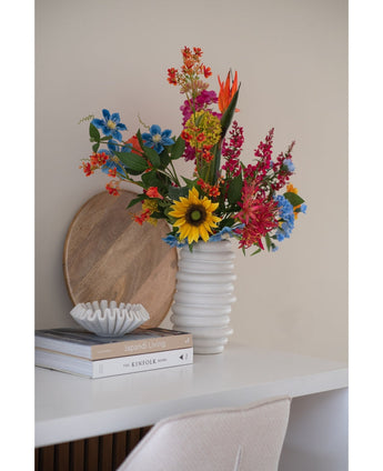 Een Kunstbloem Anjer tros 70 cm blauw in een vaas van wit keramiek staat op een wit bureau naast gestapelde boeken en een kleine decoratieve schaal, met daarachter een rond houten dienblad dat tegen de muur leunt.