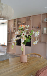 Ein modernes Esszimmer mit einem Holztisch mit einer pfirsichfarbenen Vase, in der sich die künstliche Blume Fritillary 94 cm weiß befindet. Im Hintergrund befindet sich ein großes Einbauregal, das mit stilvollen Gegenständen dekoriert ist.