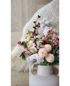 Die 70 cm große rosafarbene Kunstblume Jilly steht in einer beigen Vase auf einem Holzboden neben einem weichen elfenbeinfarbenen Schaffellteppich.