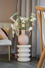 Der künstliche Blumen-Scheckenfalter 94 cm weiß steht in einer hellrosa Keramikvase mit Henkeln, platziert auf einem gewellten weißen Sockel neben einem modernen Stuhl mit gestreiftem Kissen und neutralen Vorhängen in einem stilvollen Raum.