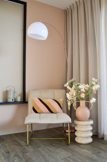 Eine moderne, gemütliche Ecke mit einem cremefarbenen Stuhl mit gestreiftem Kissen, einer hohen bogenförmigen Stehlampe, beigen Vorhängen, einem Holzboden und einer runden Vase mit weißen Blumen des künstlichen Perlmutterfalters (94 cm) auf einem geformten Beistelltisch.