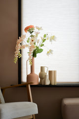 Eine pastellfarbene Wohnzimmerecke mit einem Holzstuhl, einem weichen Fußhocker und einer Fensterbank mit der 94 cm großen weißen Kunstblume „Artistic Flower Fritillary“ in einer rosa Vase neben minimalistischem Keramikdekor vor weißen Jalousien.