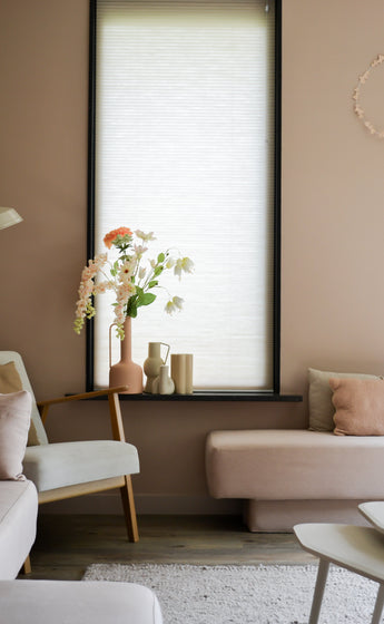 Ein gemütliches Wohnzimmer mit hellrosa Wänden, einem großen Fenster mit einem weißen Rollo, cremefarbenen und rosafarbenen Sofas und einem Holzstuhl mit der künstlichen Blumen-Perlmutterfalter 94 cm weiß neben Dekorationsgegenständen auf der Fensterbank.