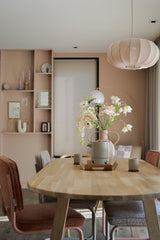 Ein modernes Esszimmer mit einem hellen Holztisch, vier Stühlen mit Kissen, dem künstlichen Perlmutterfalter 94 cm weiß in einer Vase, Keramikbechern, pastellrosa Wänden, eingebauten Regalen mit Dekor und einer großen runden Hängelampe über Ihrem Kopf.