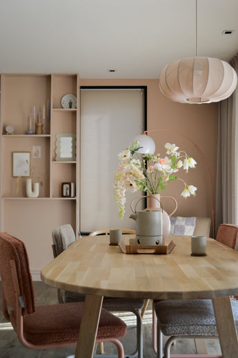 Ein modernes Esszimmer mit einem hellen Holztisch, vier Stühlen mit Kissen, dem künstlichen Perlmutterfalter 94 cm weiß in einer Vase, Keramikbechern, pastellrosa Wänden, eingebauten Regalen mit Dekor und einer großen runden Hängelampe über Ihrem Kopf.