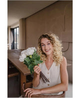 Een glimlachende vrouw met krullend blond haar in een mouwloos beige topje zit in een moderne kamer en houdt een Zijden kunstbloemen boeket Peony Wit vast. Ze kijkt naar de camera met een houten tafel zichtbaar op de achtergrond.