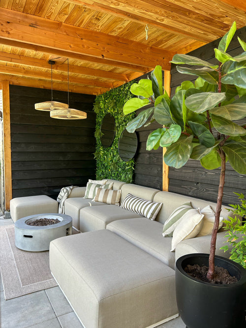 Ein gemütlicher Sitzbereich im Freien mit einem beigen Sofa, gestreiften Kissen, einer runden Feuerstelle, einer großen Topfpflanze und einer Holzpergola. Für zusätzlichen Stil wird die Wand mit dem künstlichen Haag Volta 100x100 cm UV mit Grün dekoriert.